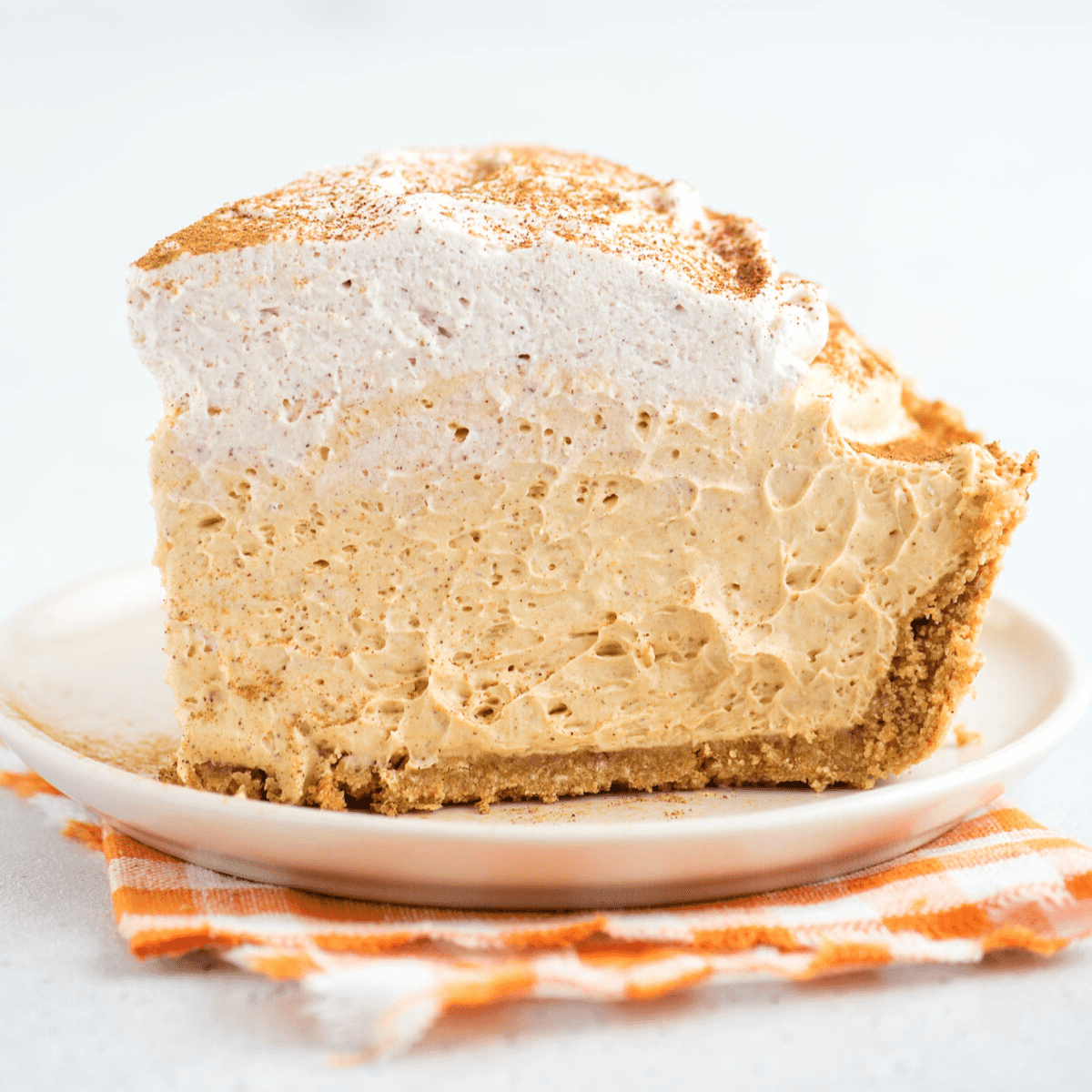 Un pezzo leggero e cremoso di torta di zucca senza cottura adagiato su un piatto da dessert, sopra un tovagliolo a quadretti arancioni.