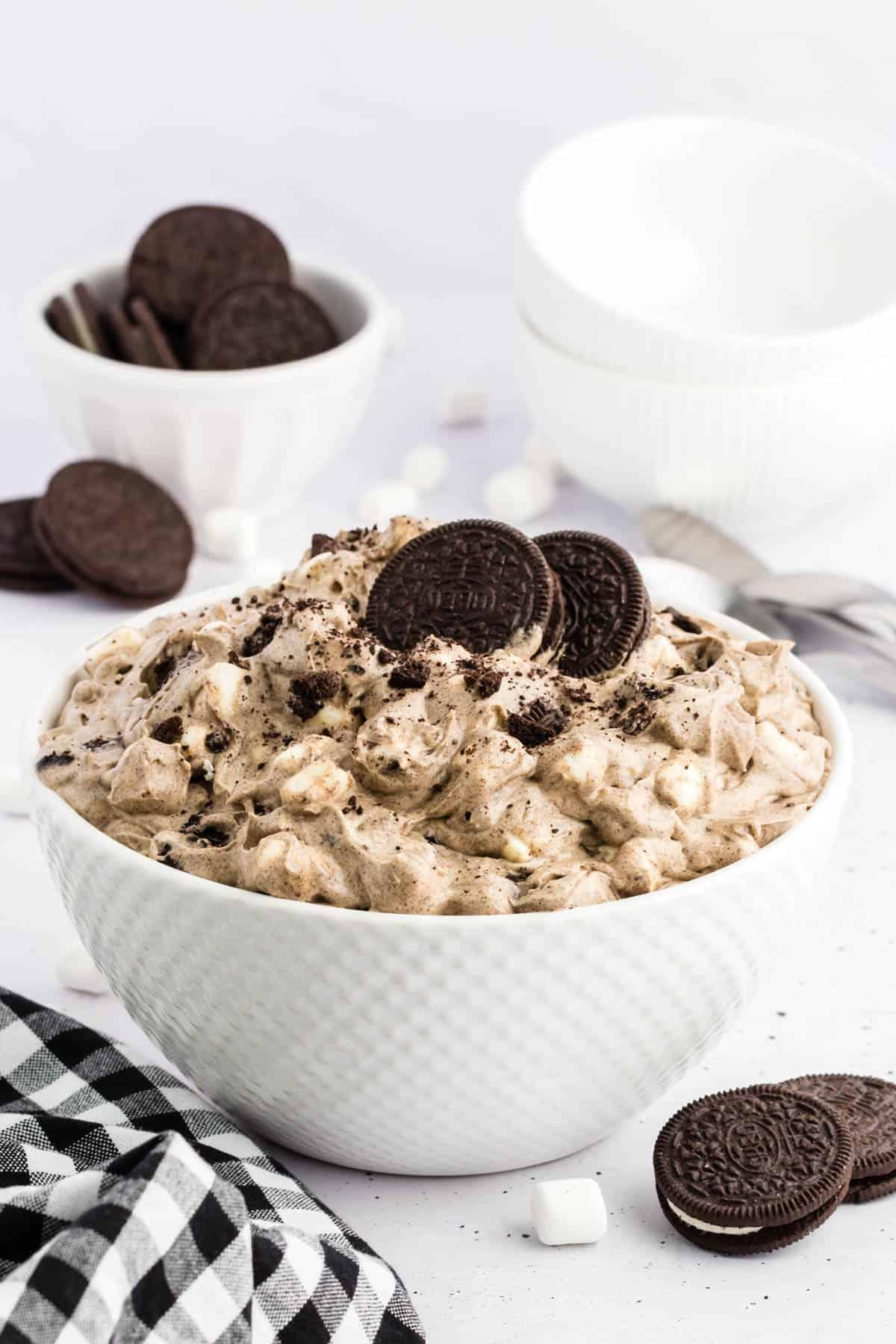 Fluff di Oreo in una ciotola bianca circondato da biscotti Oreo interi, mini marshmallow e un panno da cucina a scacchi bianchi e neri