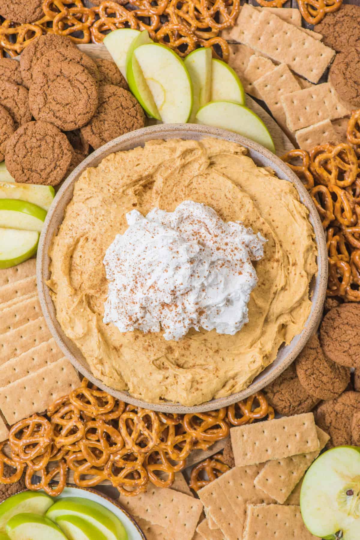 Cremosa crema di zucca con panna montata, circondata da pretzel, biscotti allo zenzero, cracker Graham e fette di mela verde su un piatto di legno.
