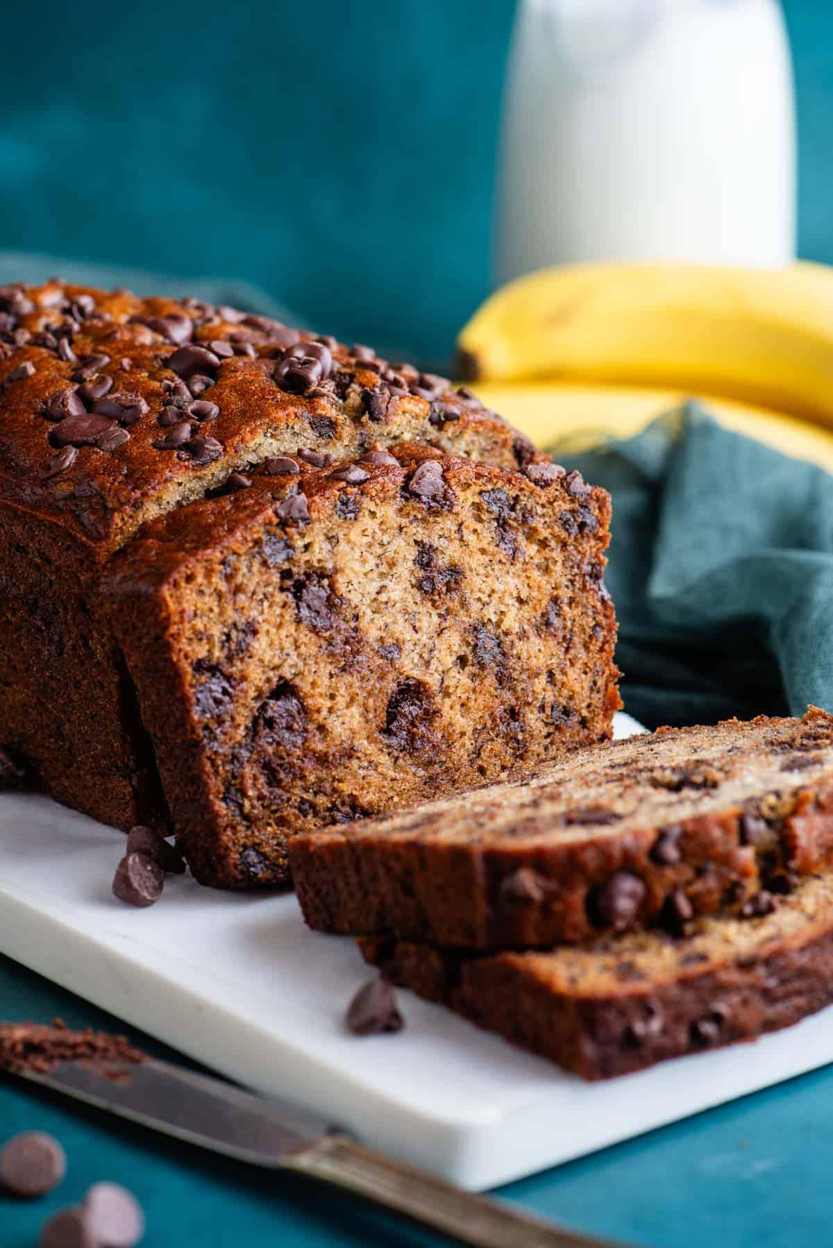 Una pagnotta di pane alle banane e gocce di cioccolato su un tagliere bianco con un asciugamano color ottanio scuro accanto, gocce di cioccolato sparse intorno, e banane intere e un bicchiere di latte sullo sfondo.