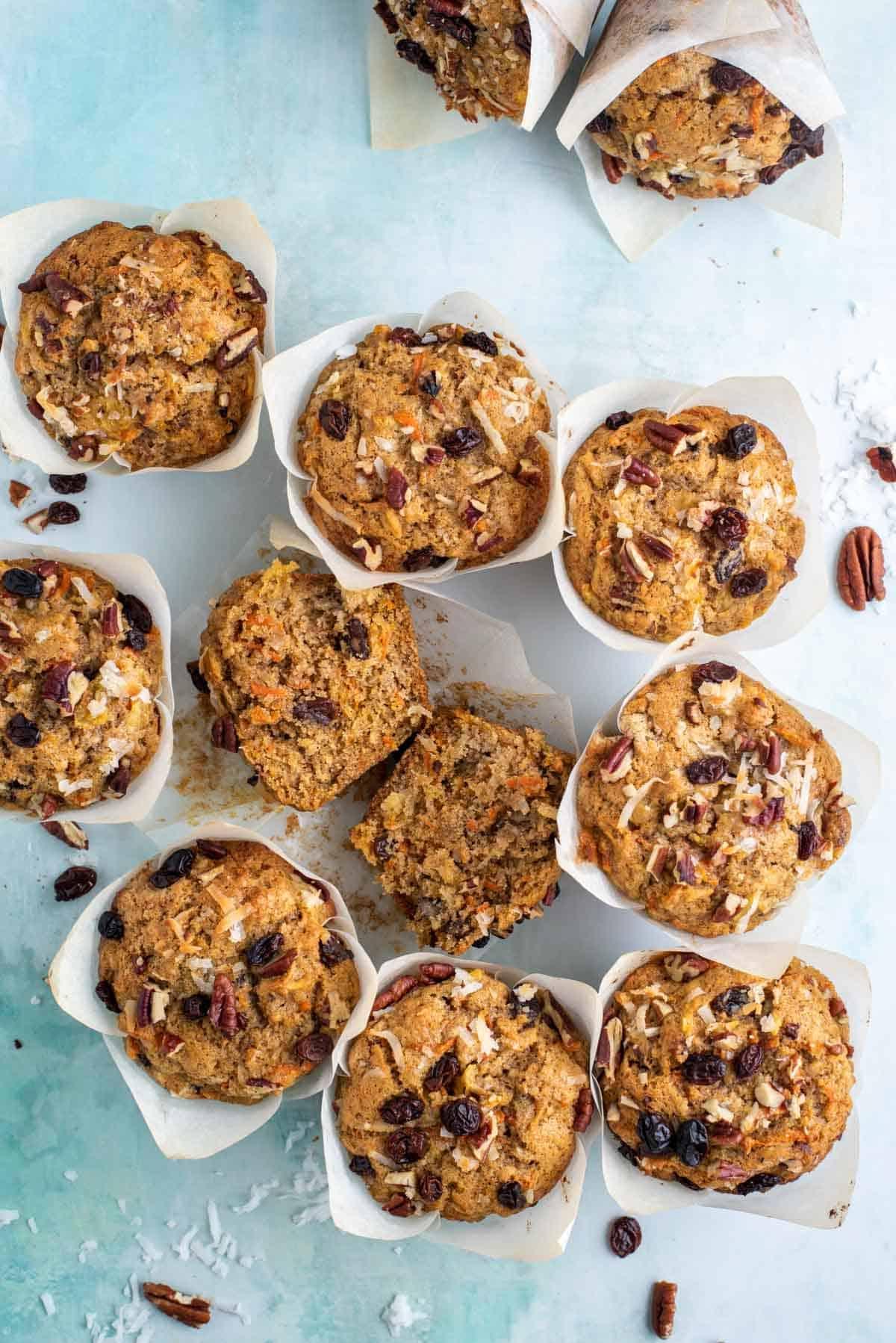 Un muffin Morning Glory tagliato a metà all'interno del suo pirottino di carta, circondato da altri muffin, noci, uvetta e scaglie di cocco.