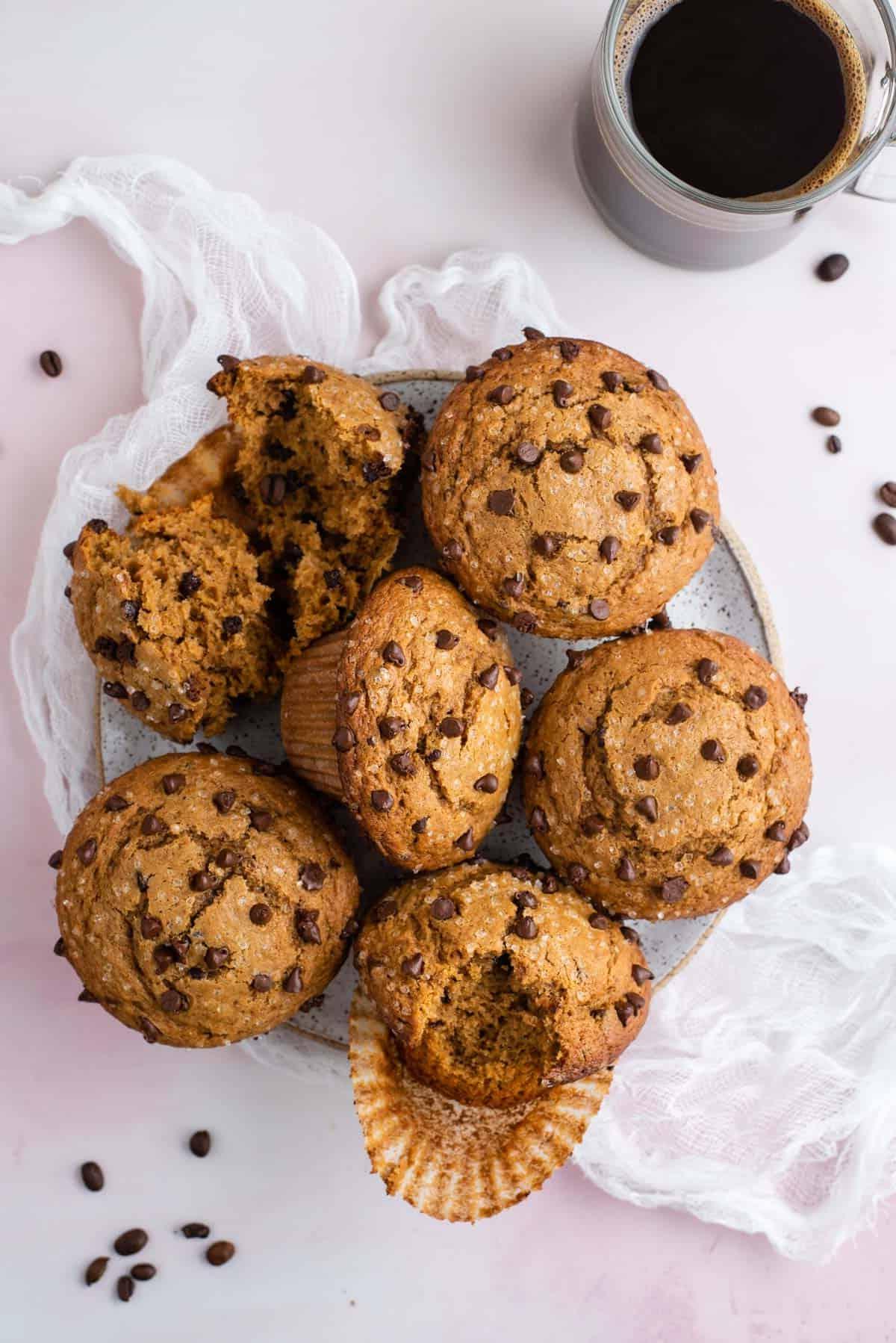 Muffin al Cappuccino e Gocce 2 un piatto pieno di muffin al cappuccino con gocce di cioccolato su un telo di formaggio bianco con chicchi di caffè sparsi intorno e una tazza di caffè di vetro trasparente accanto