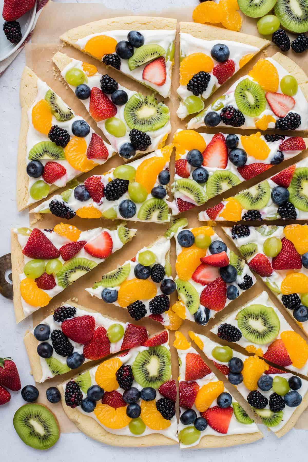 La Migliore Ricetta di Pizza alla Frutta 11 Una pizza di frutta intera tagliata a fette triangolari con fragole, mirtilli, mandarini, kiwi e uva su carta da forno marrone