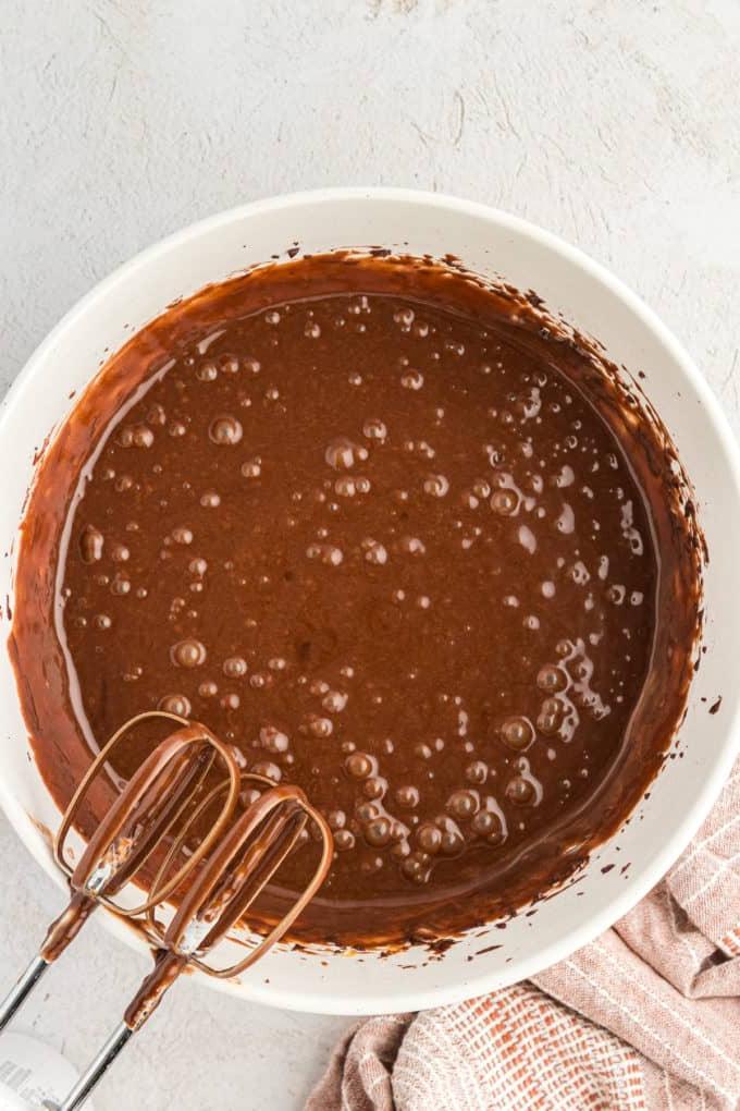 Impasto per torta Texana in una ciotola bianca con i mixer appoggiati alla ciotola coperti di impasto e un panno da cucina color cuoio a sinistra della ciotola