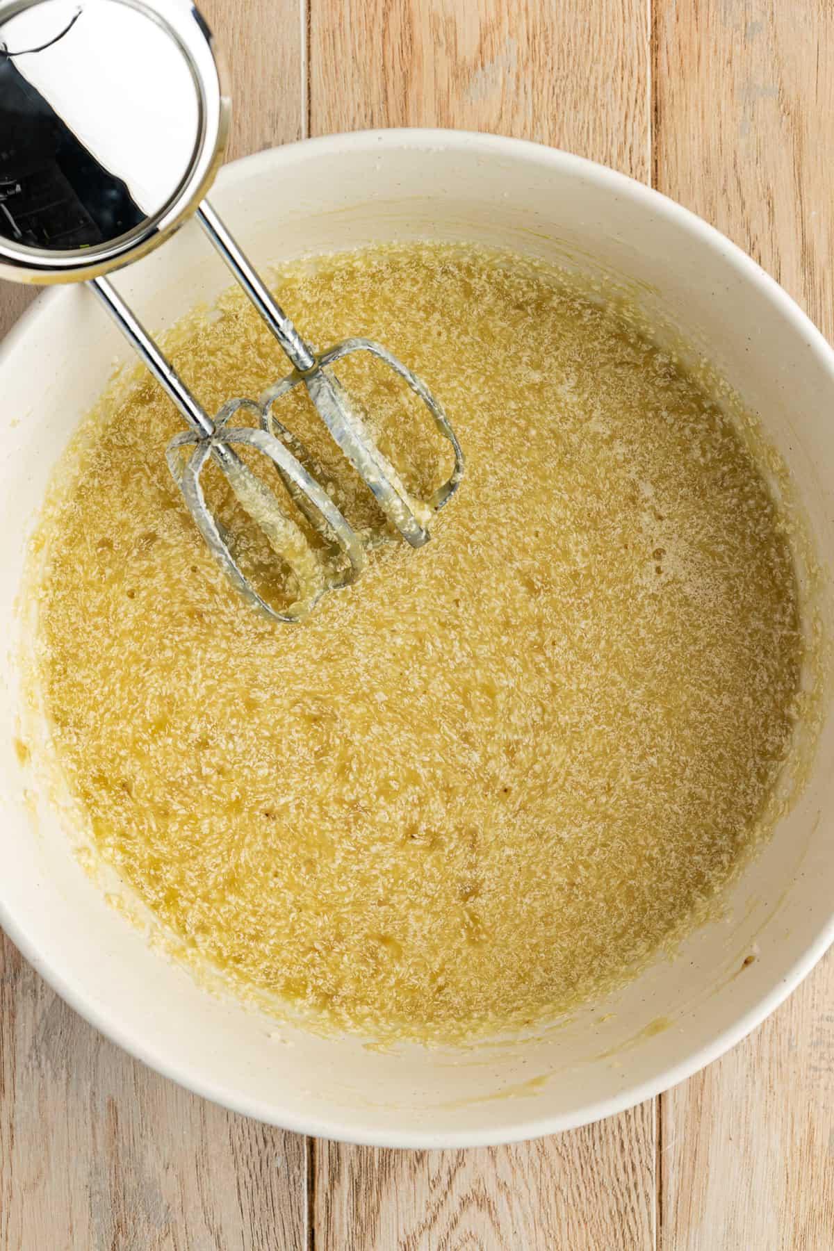 Una ciotola bianca su una superficie di legno con un impasto di torta di banane e un mixer elettrico appoggiato all'interno della ciotola
