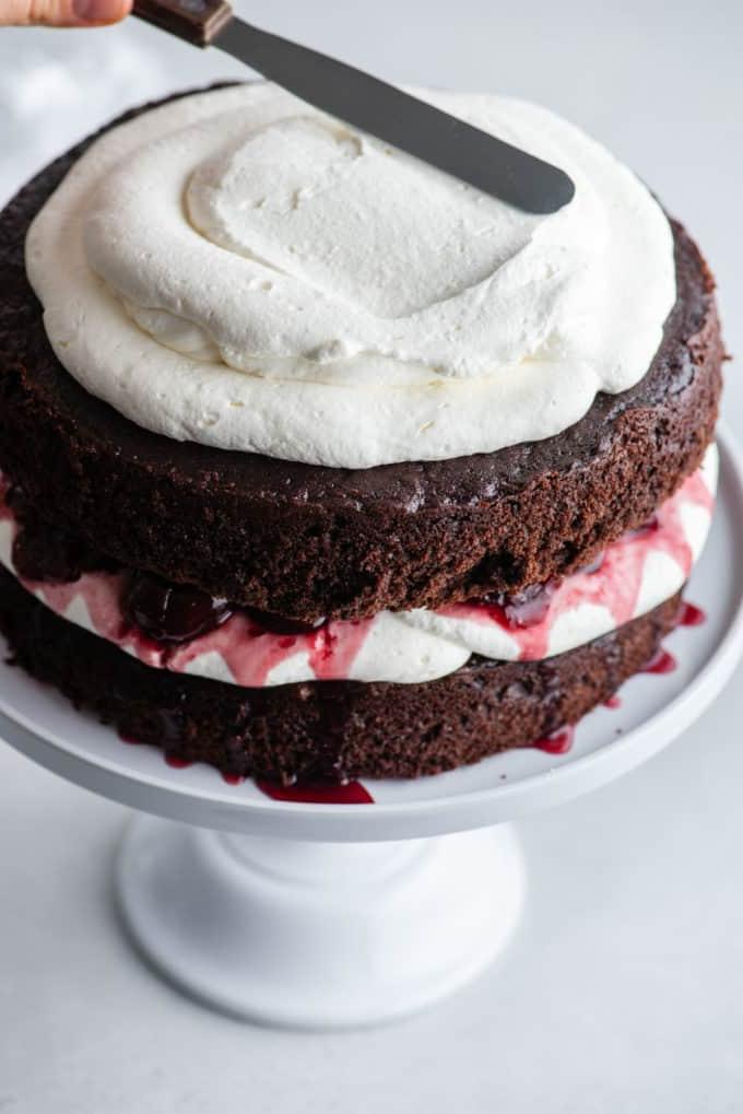 Torta Foresta Nera su un'alzatina bianca con uno strato di torta al cioccolato, panna montata, ciliegie, più torta al cioccolato e panna montata che viene spalmata sopra