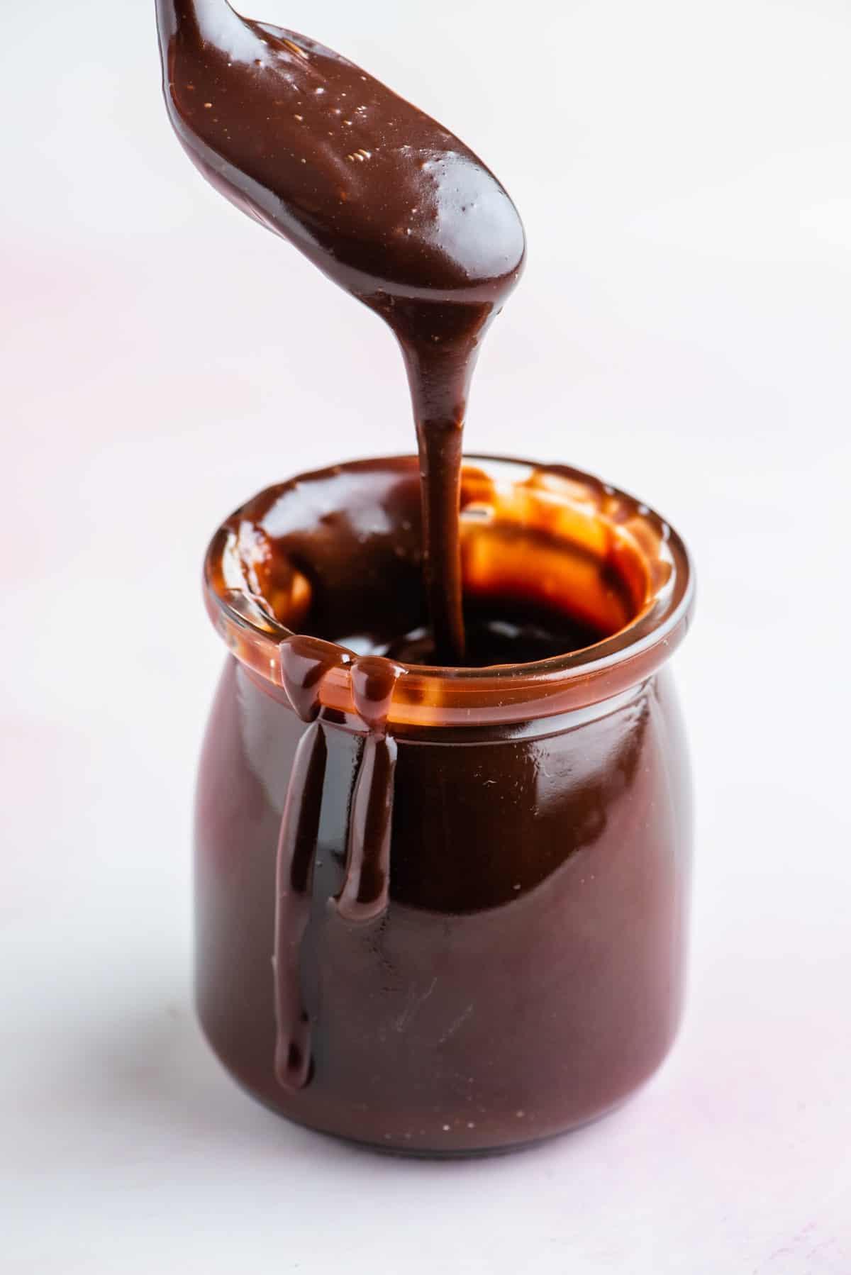 Un barattolo di salsa hot fudge con un cucchiaio sopra pieno di salsa che gocciola e salsa che gocciola lungo il lato del barattolo