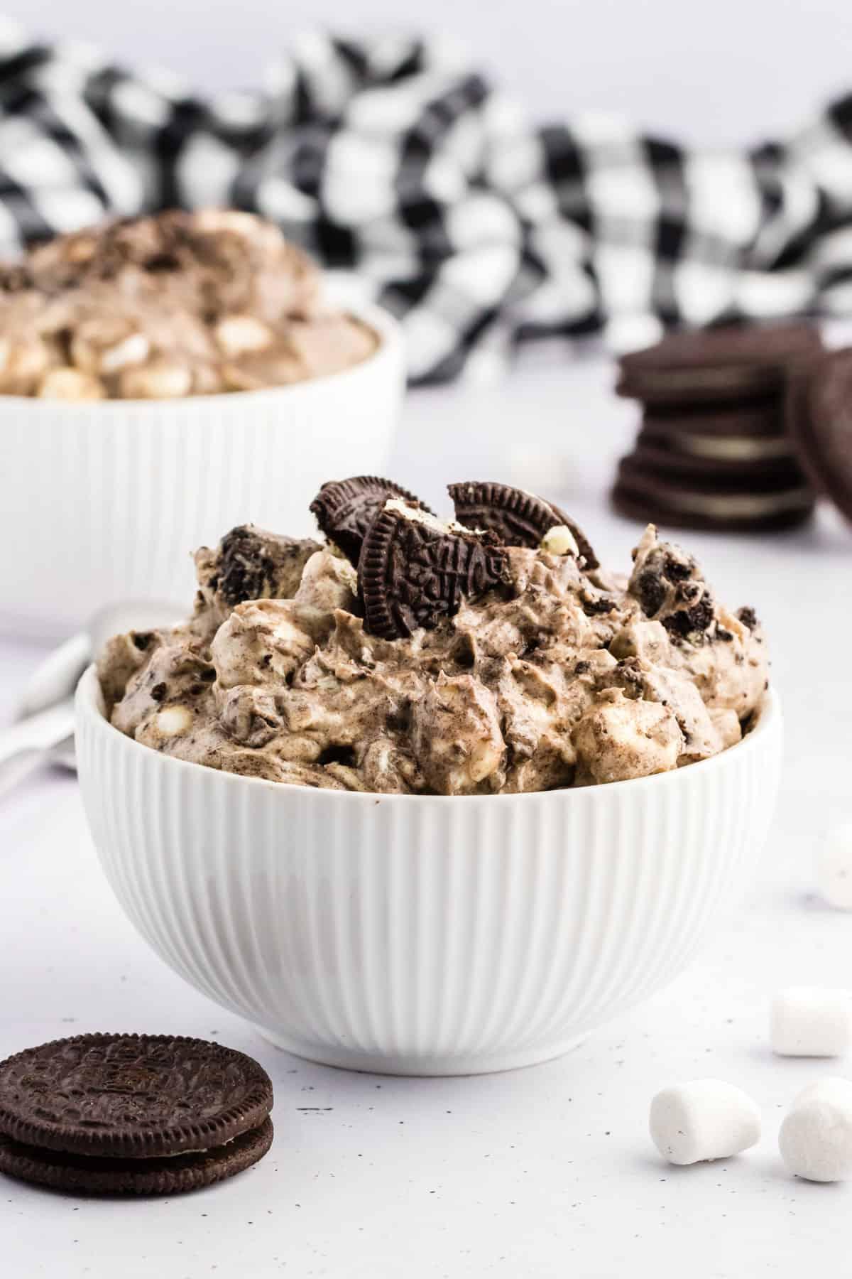 Fluff di Oreo in una ciotola bianca circondato da biscotti Oreo interi, mini marshmallow e un panno da cucina a scacchi bianchi e neri