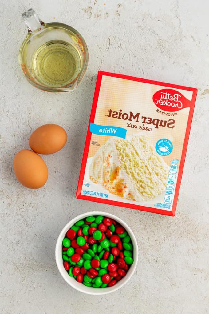 Ingredienti per biscotti natalizi con codette e red velvet su un piano di lavoro, inclusi una scatola di preparato per torte bianco, olio vegetale, due uova e una ciotola di M&M's verdi e bianchi