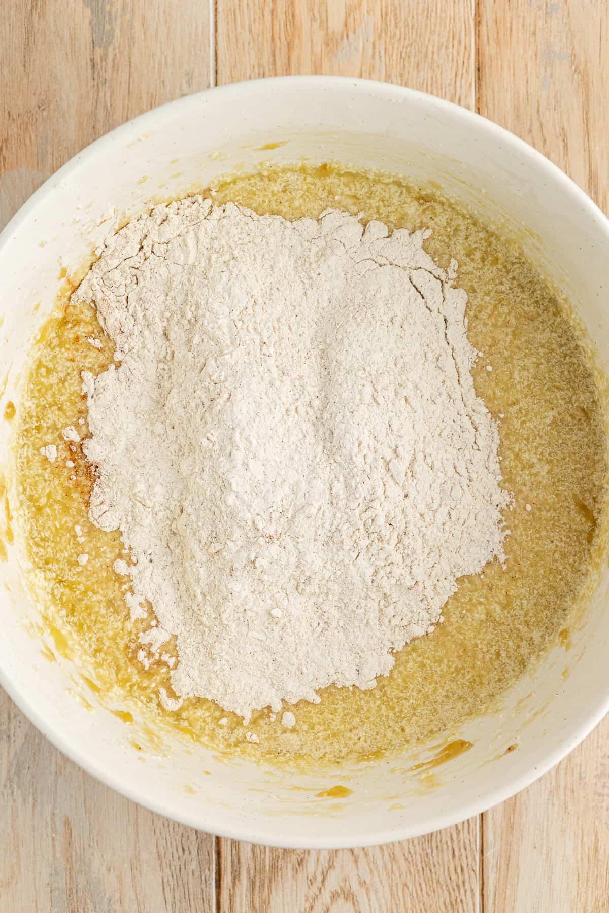 Una ciotola bianca su una superficie di legno con ingredienti umidi per la torta di banane e una miscela di farina secca sopra non ancora mescolata