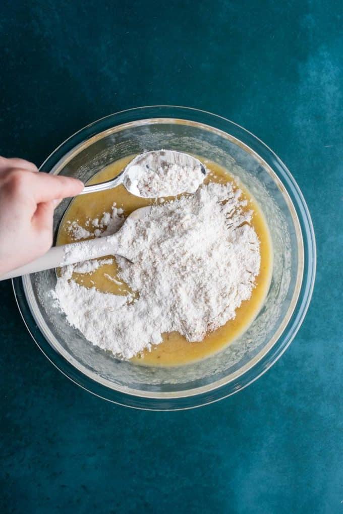 Ingredienti liquidi per il pane alle banane e gocce di cioccolato in una ciotola di vetro trasparente su una superficie color ottanio scuro con ingredienti secchi che vengono aggiunti sopra.