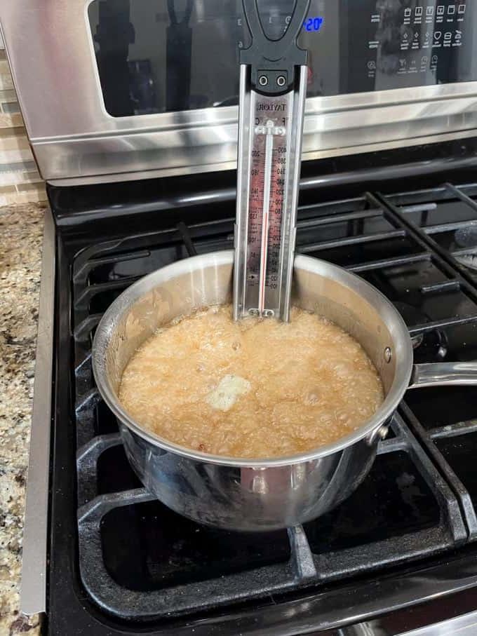 Una pentola sul fornello con zucchero semolato, acqua, sciroppo di mais, bicarbonato di sodio, estratto di vaniglia e arachidi che cuociono e un termometro per dolci attaccato al lato