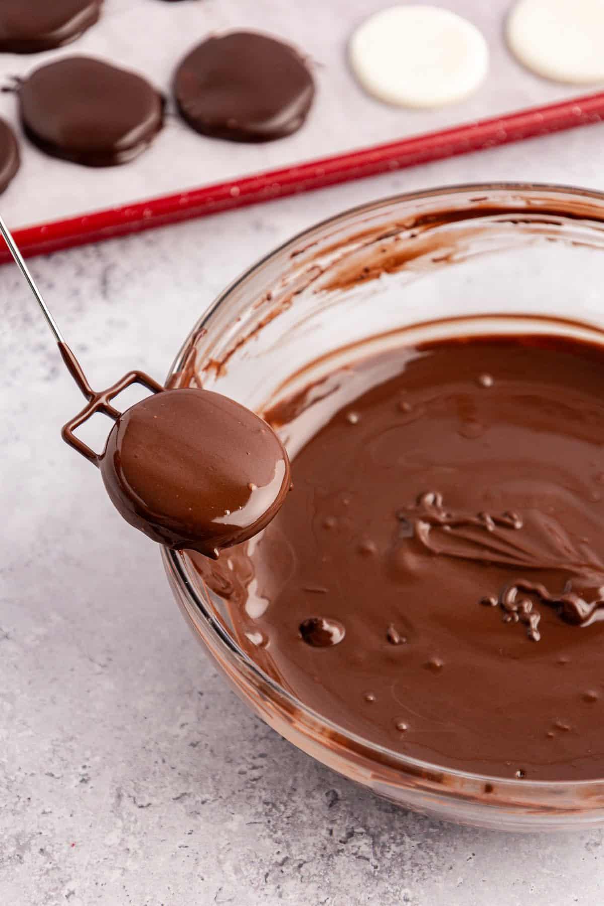Un patty alla menta appena immerso in una ciotola di vetro trasparente di cioccolato fuso viene sollevato dal bordo della ciotola