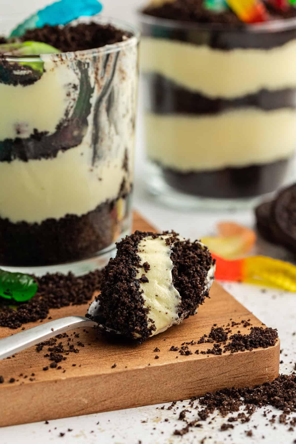 Un cucchiaio pieno di torta di terra poggiato su un piatto di legno accanto a piccole coppe da trifle di torta di terra con briciole di Oreo e vermi gommosi sparsi intorno