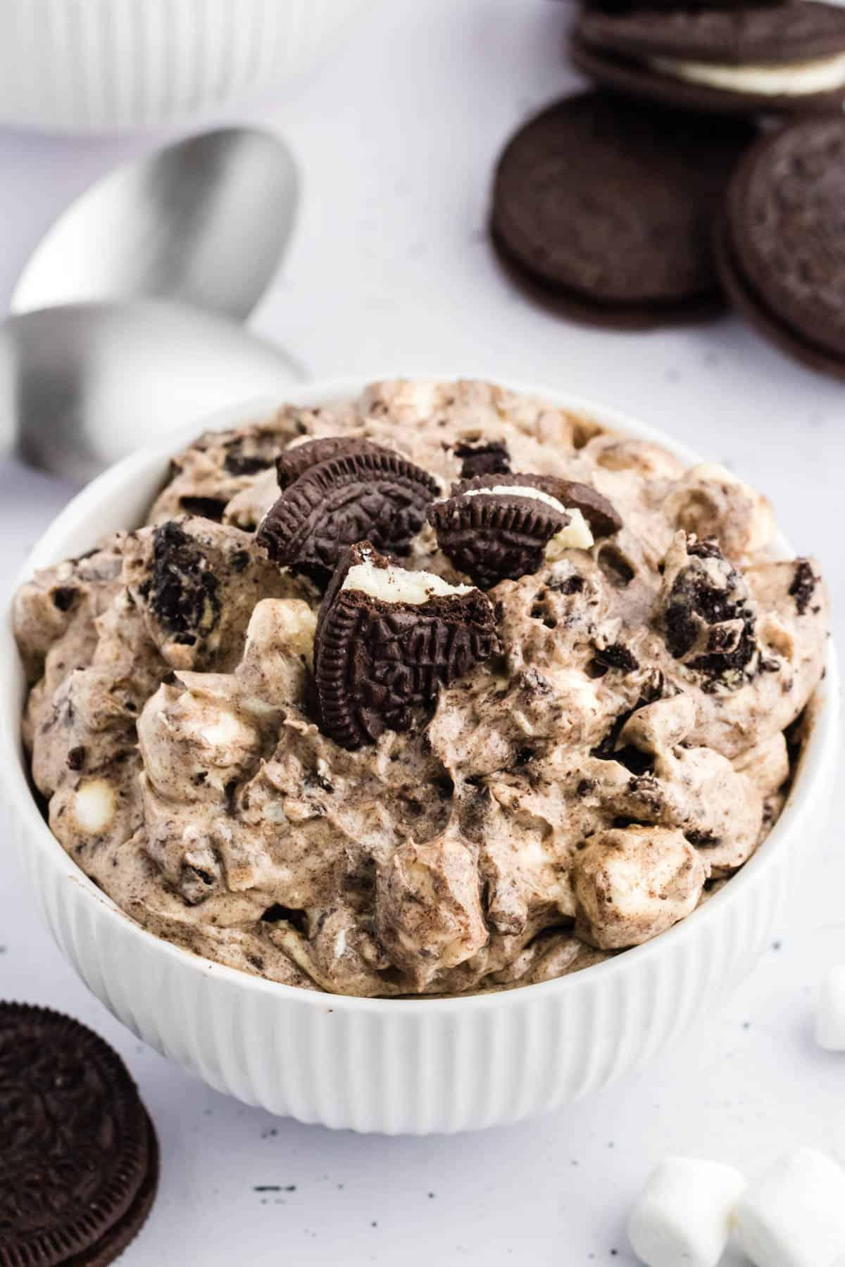 Fluff di Oreo in una ciotola bianca circondato da biscotti Oreo interi, mini marshmallow e un panno da cucina a scacchi bianchi e neri