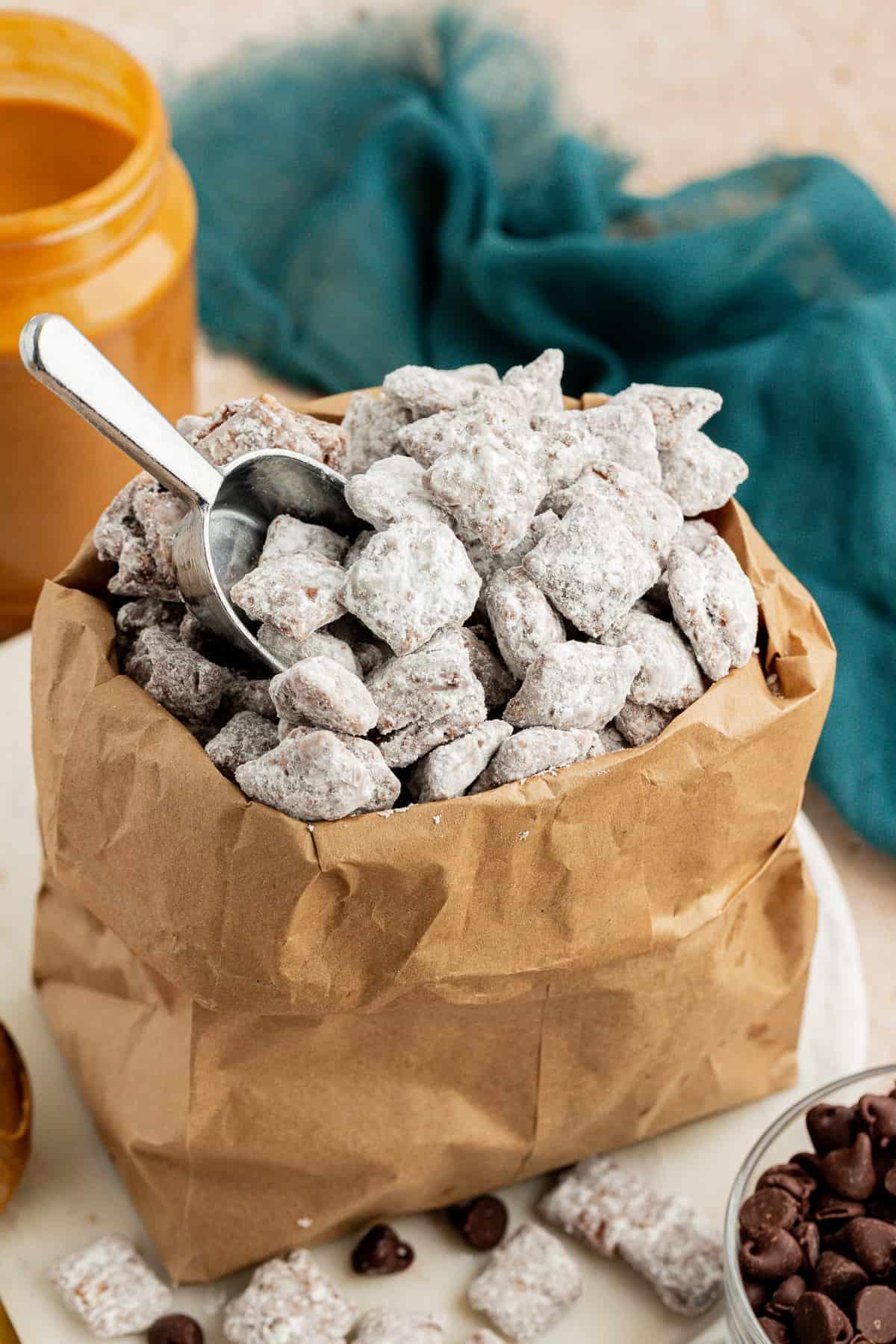 Una borsa marrone piena di puppy chow con un cucchiaio dosatore d'argento al suo interno, e un barattolo di burro d'arachidi e un asciugamano color ottanio scuro sullo sfondo e una ciotola di gocce di cioccolato accanto, con più puppy chow e gocce di cioccolato sparse intorno