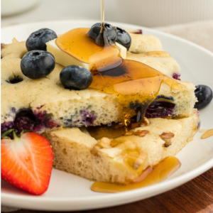 Ricetta Pancakes in Teglia 13 Due fette di pancake al forno in teglia su un piatto bianco guarnite con mirtilli freschi, una fetta di burro e sciroppo, con una fetta di fragola fresca sul piatto accanto ai pancake.