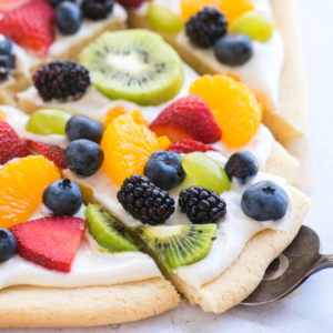 La Migliore Ricetta di Pizza alla Frutta 14 Primo piano di una deliziosa fetta di pizza di frutta guarnita con glassa al formaggio cremoso, mirtilli, more, kiwi, fragole, uva verde e spicchi d'arancia su una morbida base di biscotto zuccherato.