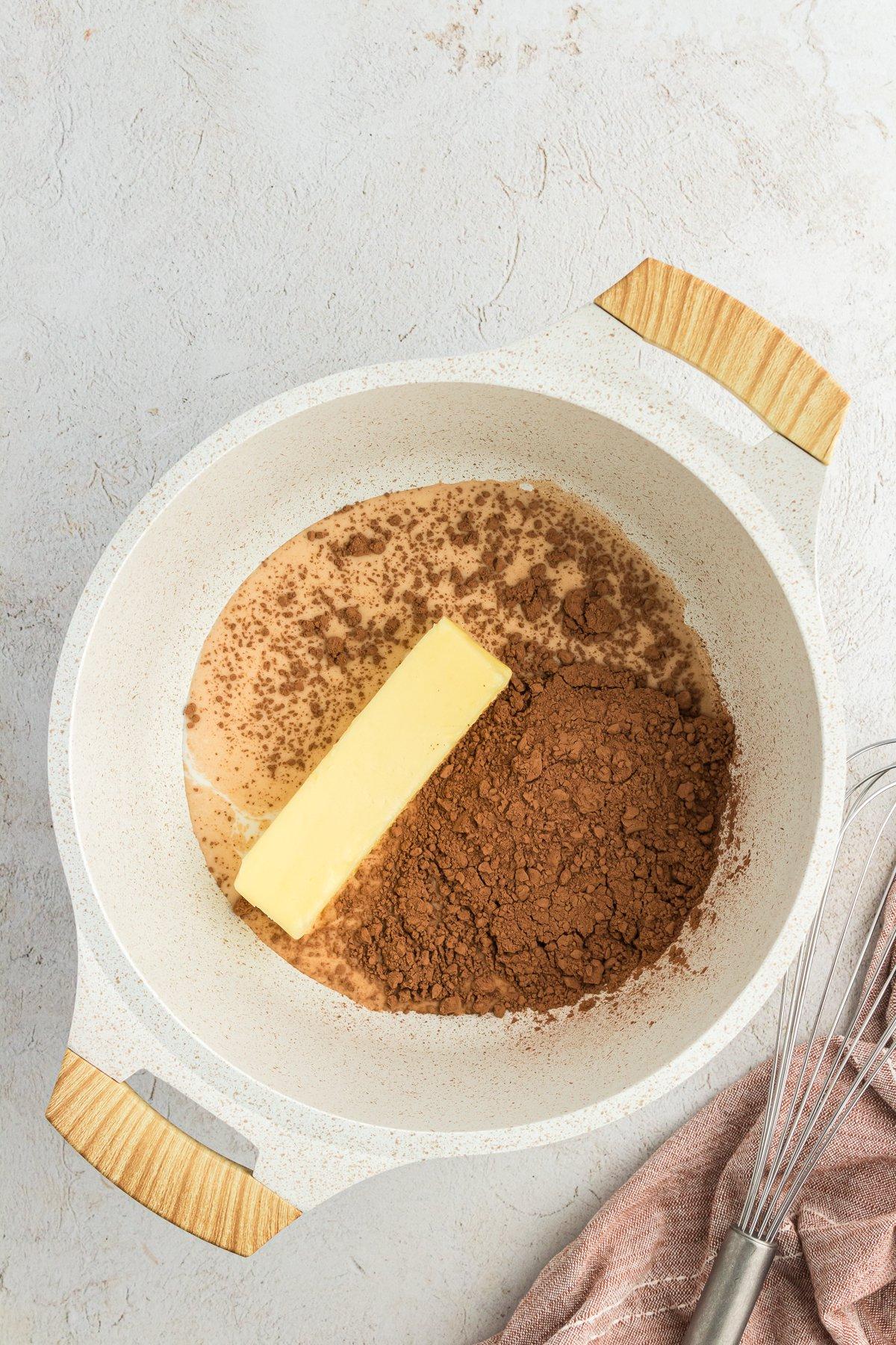 Latte, cacao in polvere e burro in una casseruola con una frusta e un panno da cucina color cuoio accanto