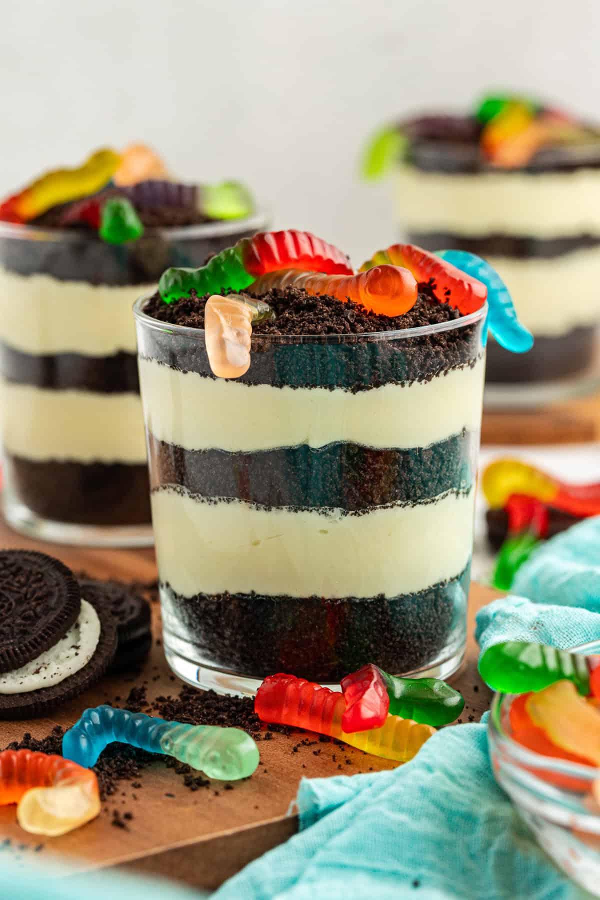 Tre piccole coppe da trifle di torta di terra guarnite con vermi gommosi su un piatto di legno circondate da Oreo, Oreo sbriciolati, vermi gommosi e un asciugamano color ottanio chiaro