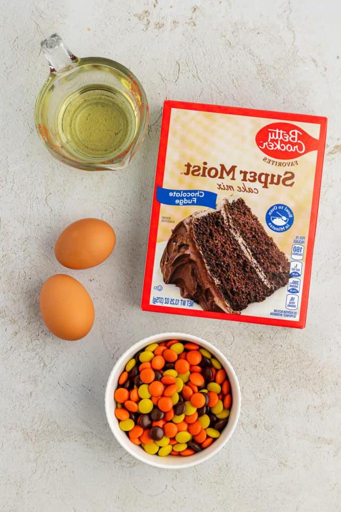 Ingredienti per biscotti con preparato per torte Reese's Pieces, inclusi una scatola di preparato per torte al cioccolato, due uova, olio vegetale e una ciotola di Reese's Pieces