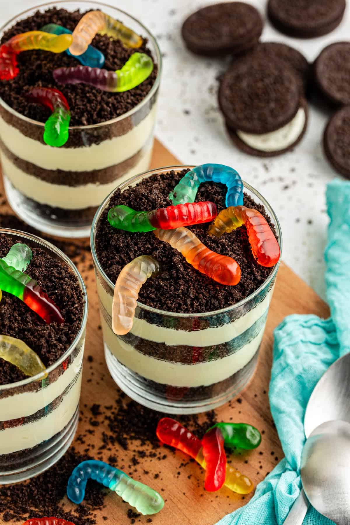 Tre piccole coppe da trifle di torta di terra guarnite con vermi gommosi su un piatto di legno circondate da Oreo interi, briciole di Oreo, vermi gommosi, cucchiai e un asciugamano color ottanio chiaro