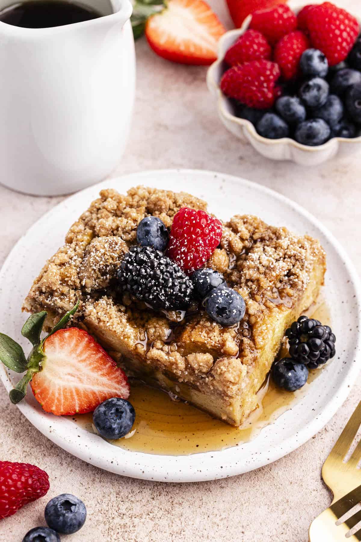 Un piattino con una fetta di casseruola di French Toast guarnita con sciroppo d'acero, frutti di bosco freschi e una leggera spolverata di zucchero a velo accanto a una piccola ciotola di frutti di bosco freschi, un piatto bianco di sciroppo, due forchette e altri frutti di bosco sparsi intorno.