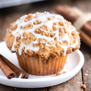 Un muffin al caffè con crumble sbriciolato e glassa bianca a filo è appoggiato su un piatto bianco, con due bastoncini di cannella accanto su una superficie di legno rustica.