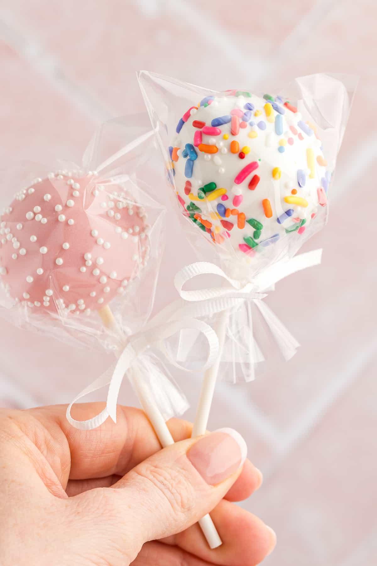 una mano che tiene due cake pops, uno bianco con codette arcobaleno e uno rosa con codette bianche, entrambi incartati nella plastica e legati con un nastro bianco