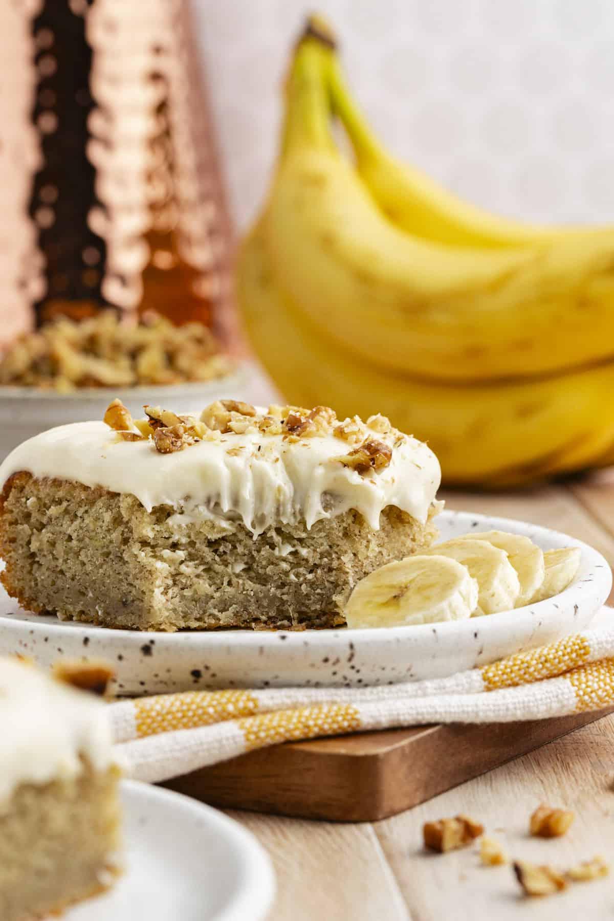 Una fetta di torta di banane su un piccolo piatto su un canovaccio a strisce bianche e gialle su un tagliere di legno, circondata da altra torta, una ciotola di noci e banane intere