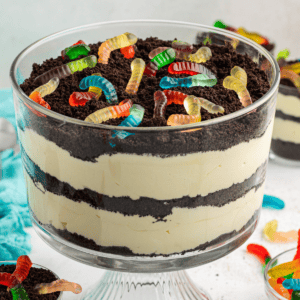 Una grande coppa da trifle di torta di terra guarnita con vermi gommosi circondata da piatti più piccoli di torta di terra, vermi gommosi e un asciugamano color ottanio chiaro