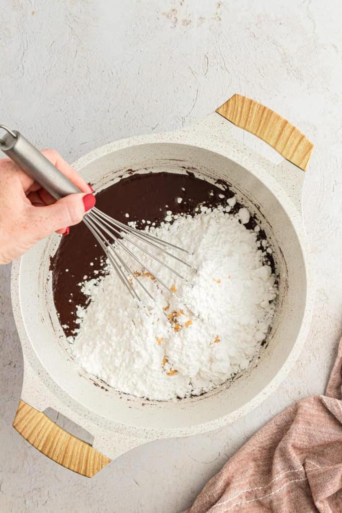 Zucchero a velo mescolato in una casseruola con glassa al cioccolato e un panno da cucina color cuoio appoggiato a sinistra della padella