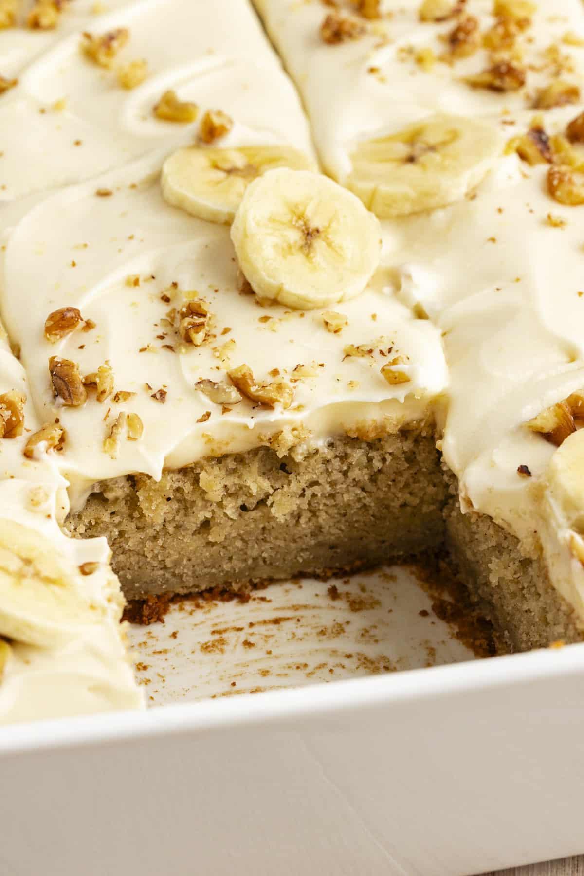 Primo piano di una teglia bianca piena di torta di banane con glassa al formaggio cremoso, con una fetta mancante che mostra l'interno con lo strato di torta e lo strato di glassa sopra