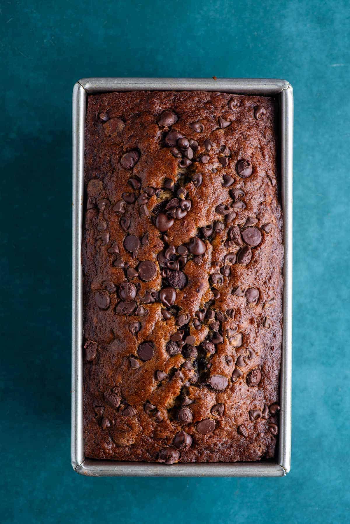 Pane alle banane e gocce di cioccolato che cuoce in uno stampo da plumcake su una superficie color ottanio scuro.