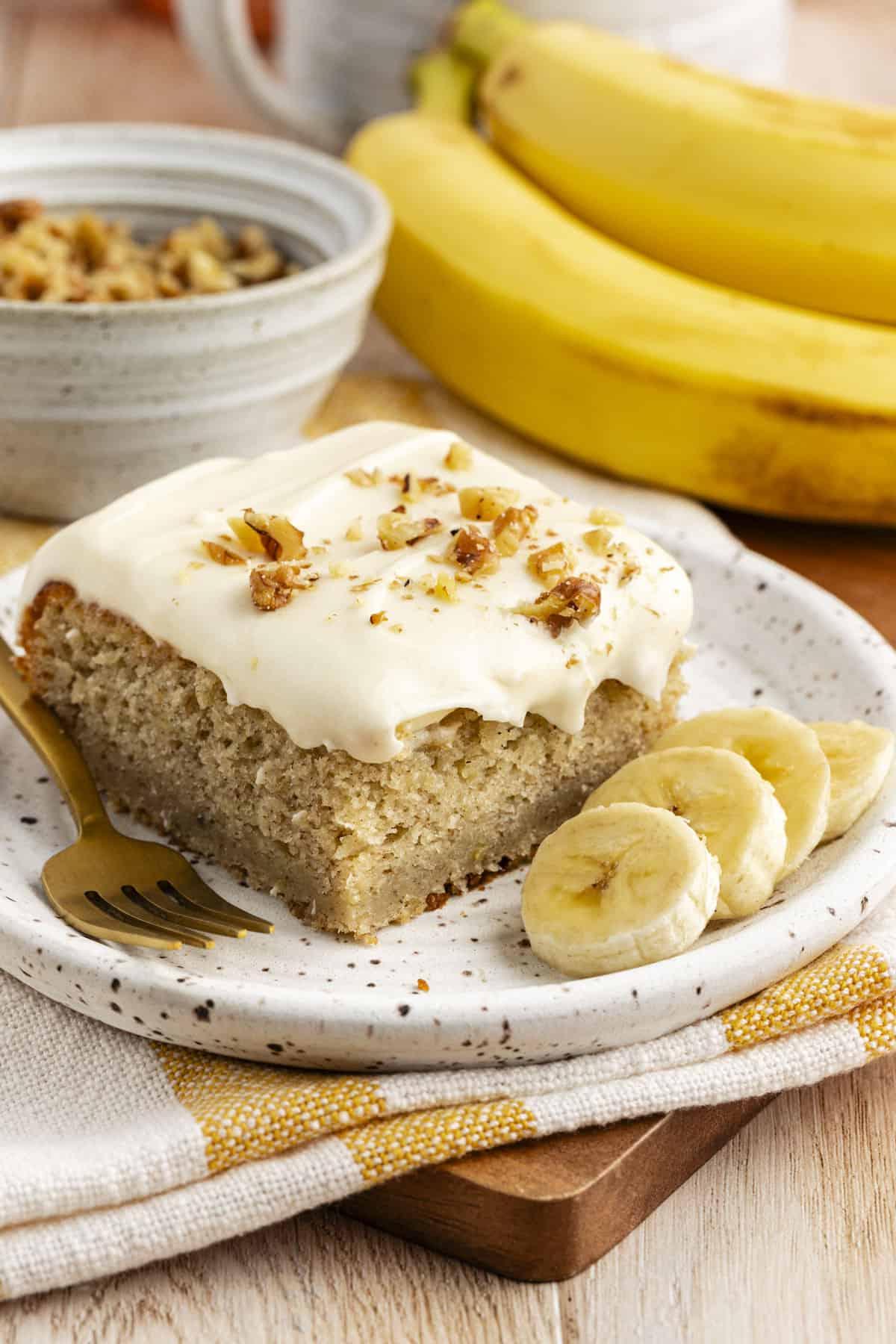 Una fetta di torta di banane con un morso mancante su un piccolo piatto con fette di banana fresca, su un canovaccio da cucina a strisce gialle e bianche con banane intere e una ciotola di crumble in sottofondo