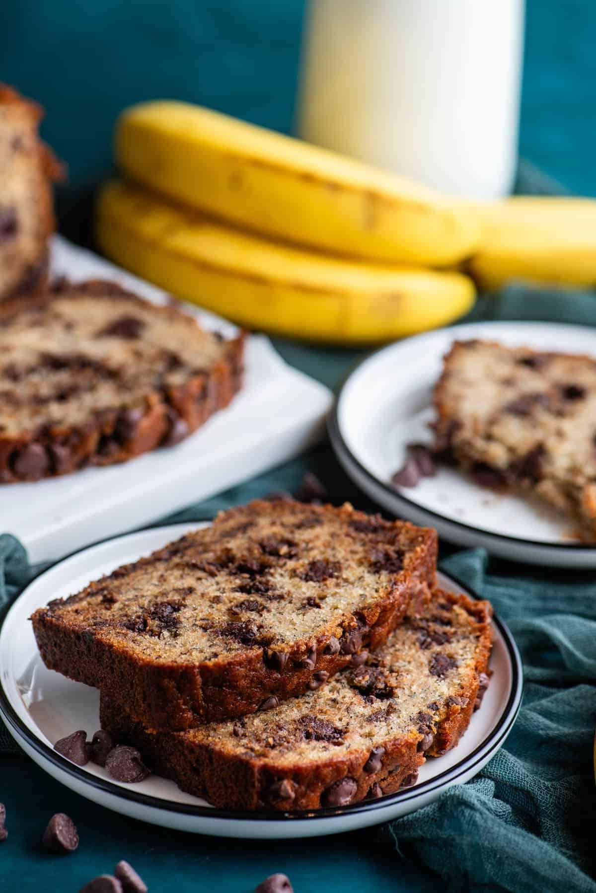 Due fette di pane alle banane e gocce di cioccolato su un piatto bianco sopra un asciugamano color ottanio scuro, con banane intere accanto, un altro piatto con una fetta di pane alle banane, e un tagliere bianco con altro pane dietro.
