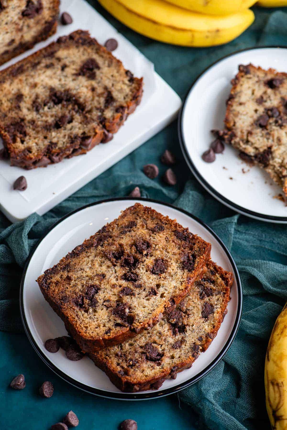 Due piccoli piatti bianchi con fette di pane alle banane, quello anteriore con due fette, e un tagliere bianco con fette di pane alle banane, il tutto appoggiato su un asciugamano color ottanio scuro con gocce di cioccolato sparse intorno.