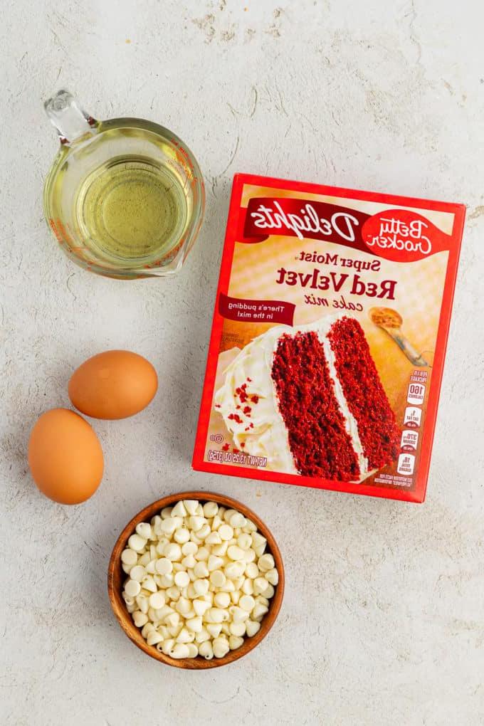 Ingredienti per biscotti Red Velvet con preparato per torte, inclusi una scatola di preparato per torte Red Velvet, due uova, una ciotola di legno di gocce di cioccolato bianco e olio vegetale