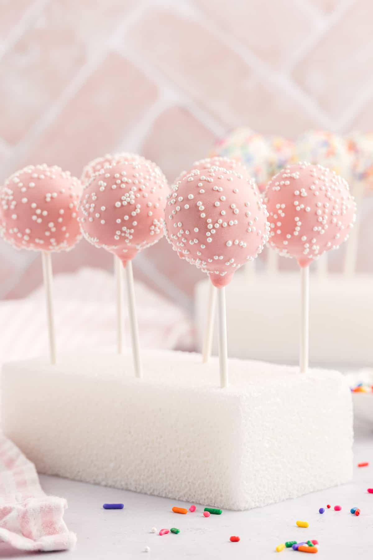 un blocco di polistirolo con 6 cake pops rosa con codette bianche infilzati per farli stare dritti, con un asciugamano a strisce bianche e rosa e codette arcobaleno sparse intorno