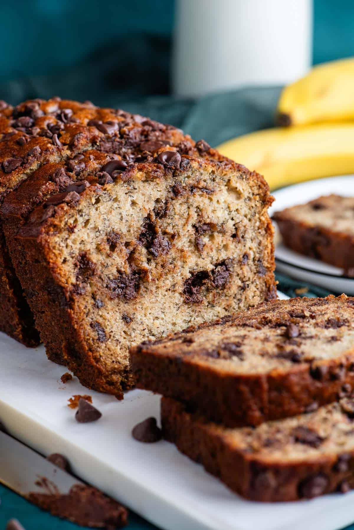 Pane alle banane e gocce di cioccolato affettato su un tagliere bianco con un piatto sullo sfondo con una fetta e banane intere dietro.