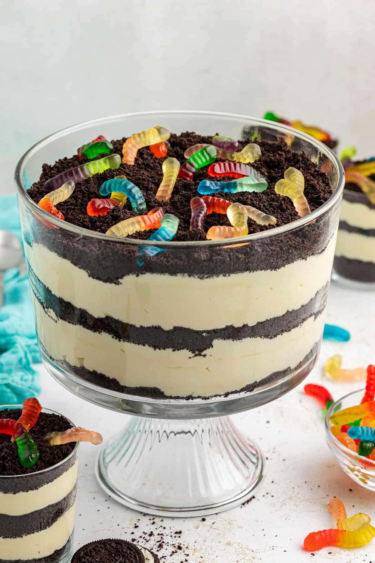 Una grande coppa di vetro trasparente da trifle piena di torta di terra guarnita con vermi gommosi, circondata da coppe più piccole di torta di terra, un asciugamano color ottanio, vermi gommosi e Oreo