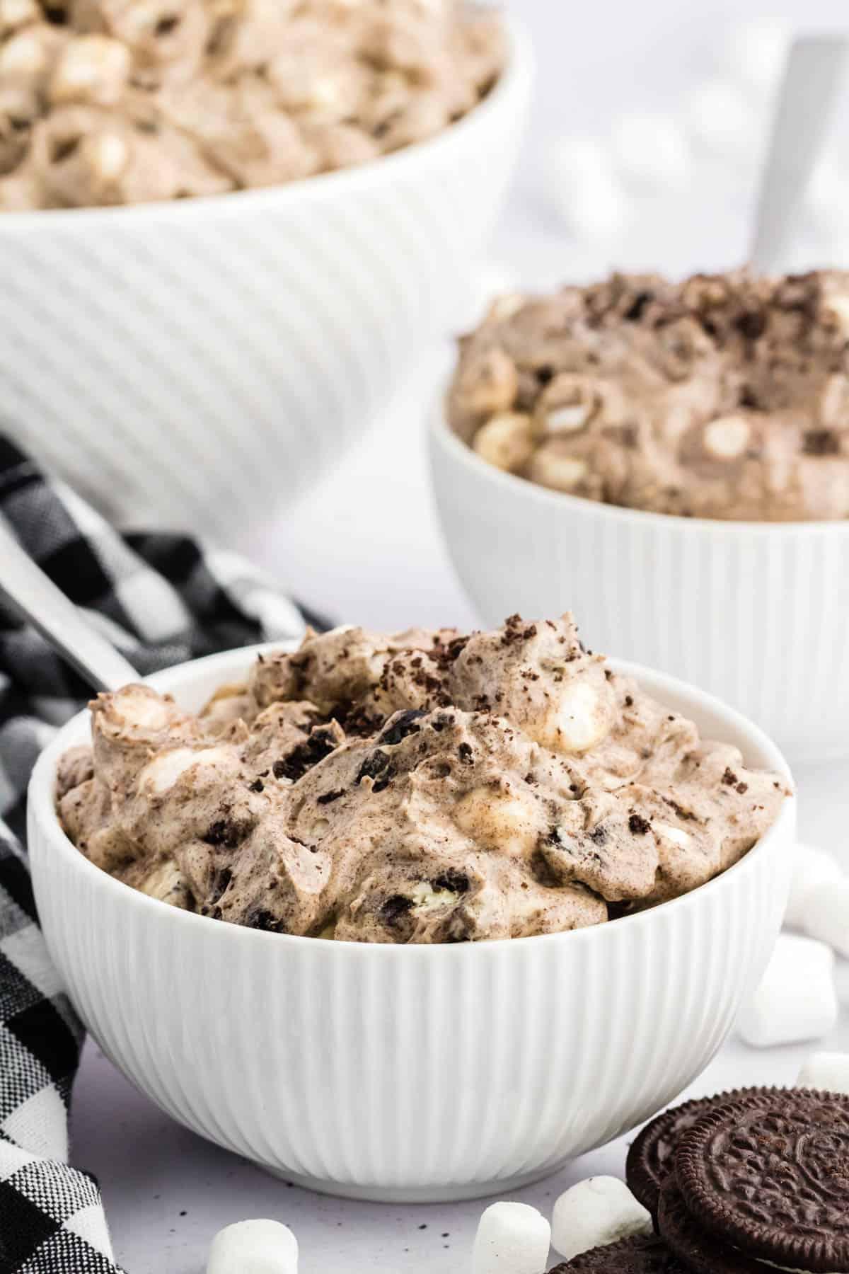 3 ciotole di fluff di Oreo circondate da biscotti Oreo interi, mini marshmallow e un panno da cucina a scacchi bianchi e neri