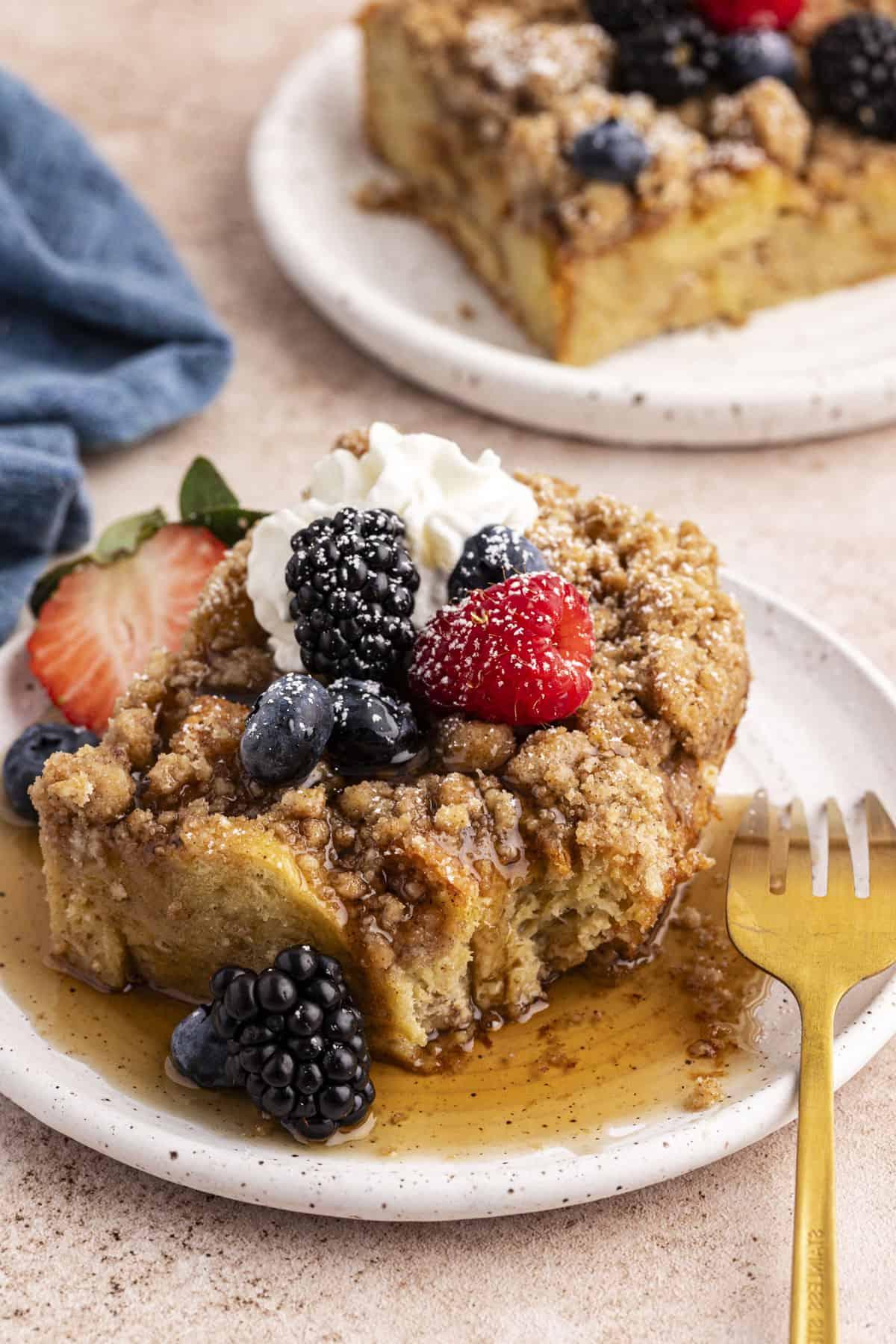 Due piattini con una fetta di casseruola di French Toast ciascuno, coperti di sciroppo d'acero, frutti di bosco freschi, un ciuffo di panna montata e una leggera spolverata di zucchero a velo, con il piatto anteriore che ha una forchetta appoggiata e un canovaccio blu sullo sfondo.