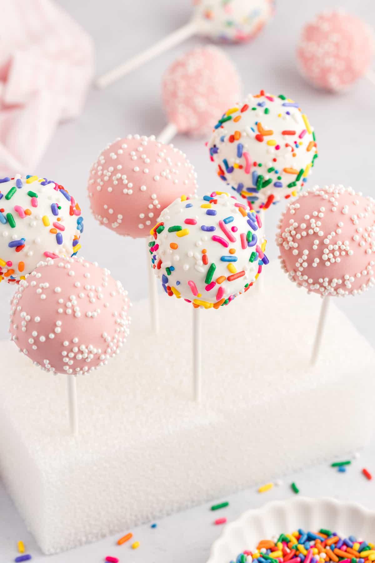 un blocco di polistirolo con sei cake pops infilzati, tre rosa con codette bianche e tre bianchi con codette arcobaleno, con una ciotola bianca di codette arcobaleno in primo piano e altri cake pops adagiati sullo sfondo