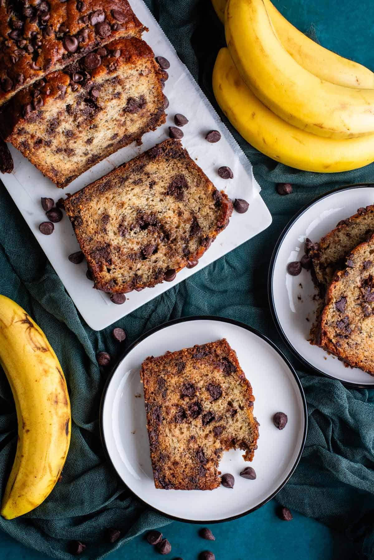 Vista dall'alto di due piatti bianchi e un tagliere bianco con fette di pane alle banane e gocce di cioccolato, tutti appoggiati su un asciugamano color ottanio scuro con gocce di cioccolato e banane intere sparse intorno.