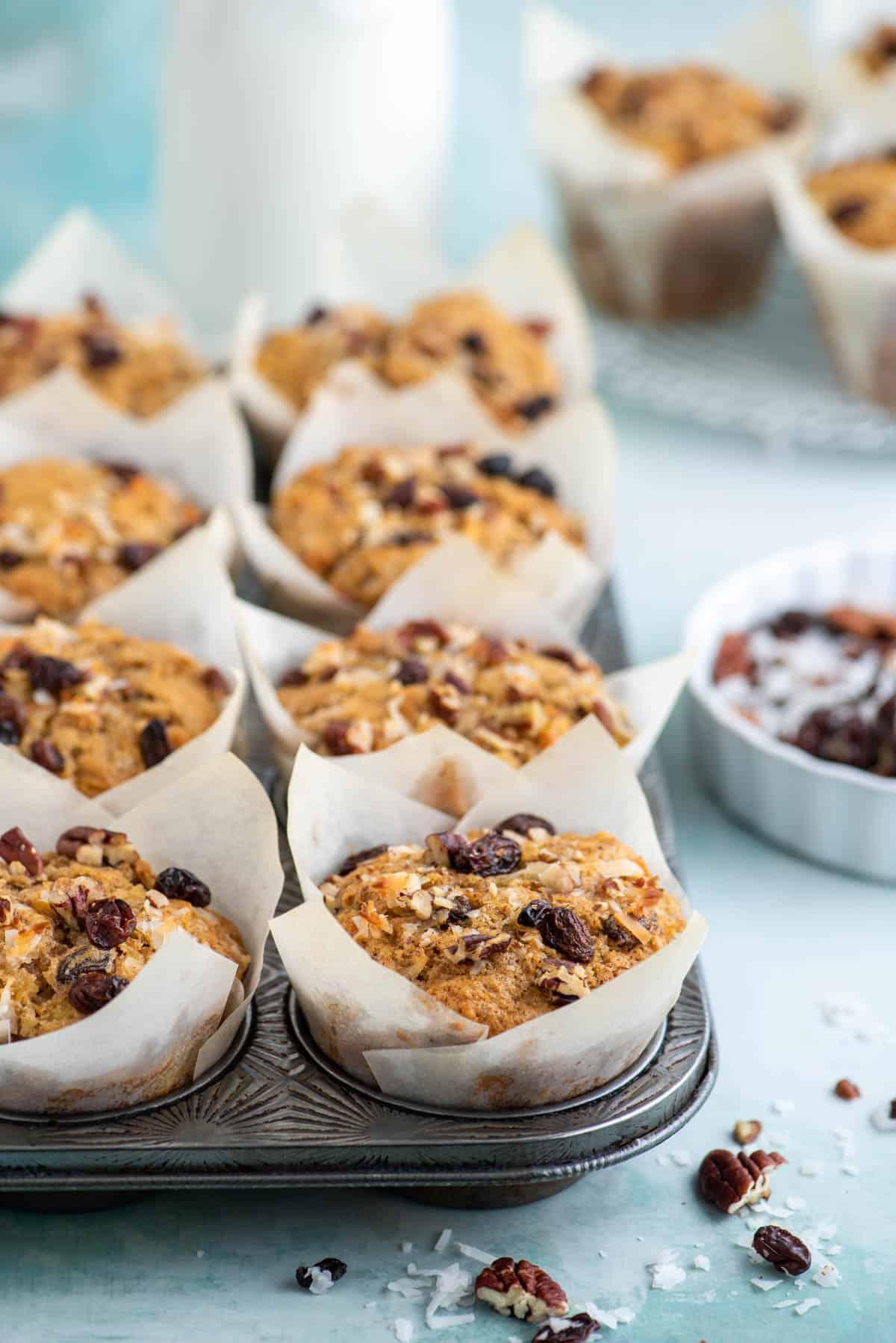 Muffin Morning Glory nei pirottini di carta all'interno di una teglia per muffin, circondati da noci, uvetta e scaglie di cocco, con altri muffin sullo sfondo.