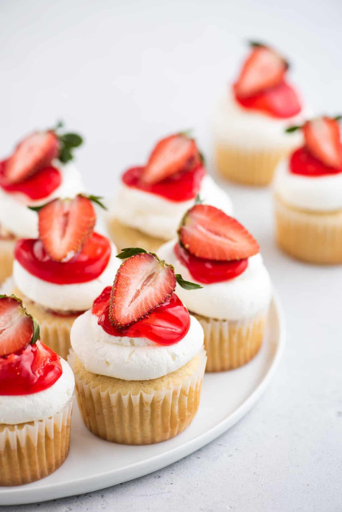 I Cupcake alle Fragole Shortcake sono fatti con torta alla vaniglia umida, ripieni di dolce salsa di fragole e ricoperti di soffice panna montata! Cupcake alle fragole shortcake su piatto rotondo e tavolo