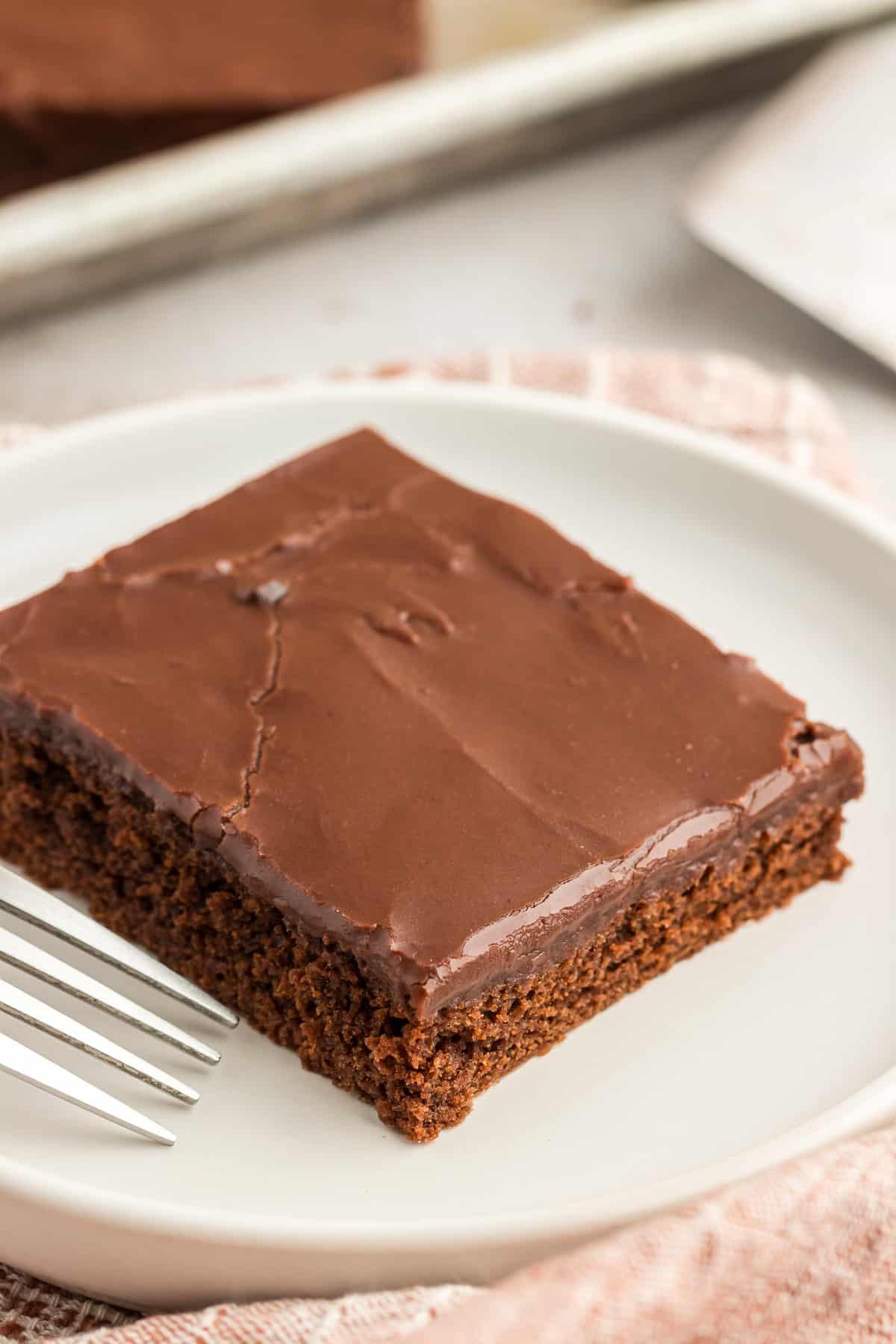 Una fetta di torta Texana al cioccolato su un piatto bianco con una forchetta e un panno da cucina color cuoio accanto