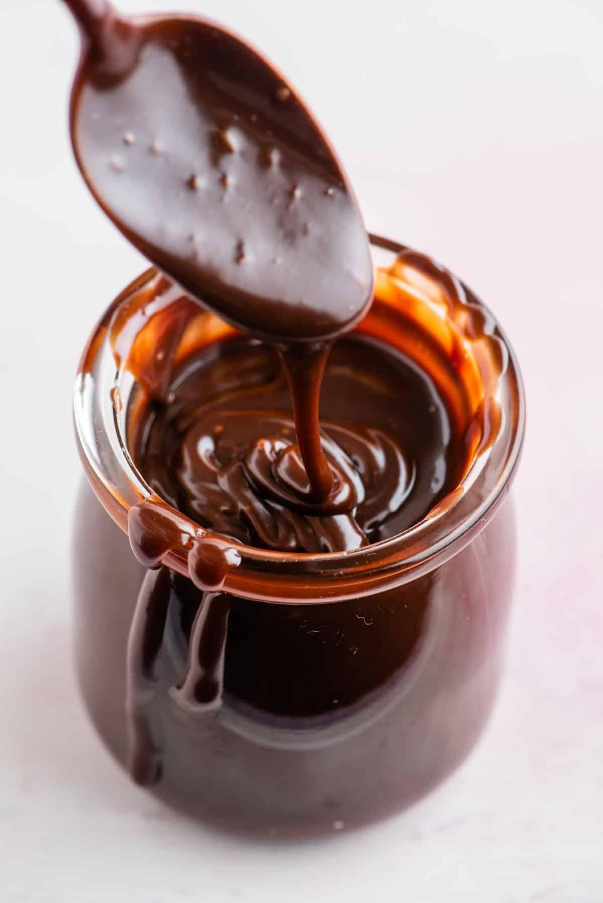 Ricetta salsa hot fudge fatta in casa in un barattolo di vetro con un cucchiaio sopra il barattolo che gocciola un po' di salsa nel barattolo