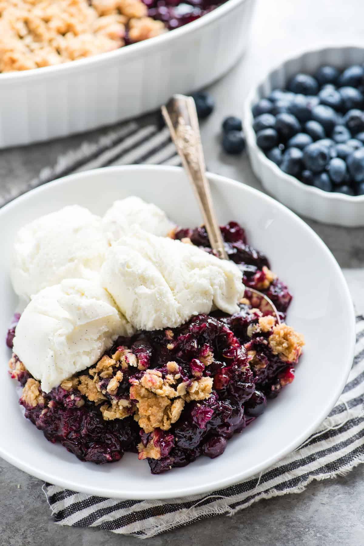 Crumble ai mirtilli in una ciotola bianca con gelato alla vaniglia e un cucchiaio di metallo su un tovagliolo a righe blu